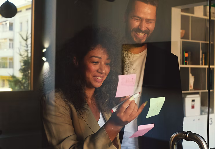 Two colleagues happily collaborate together, placing post-its on a glass window.