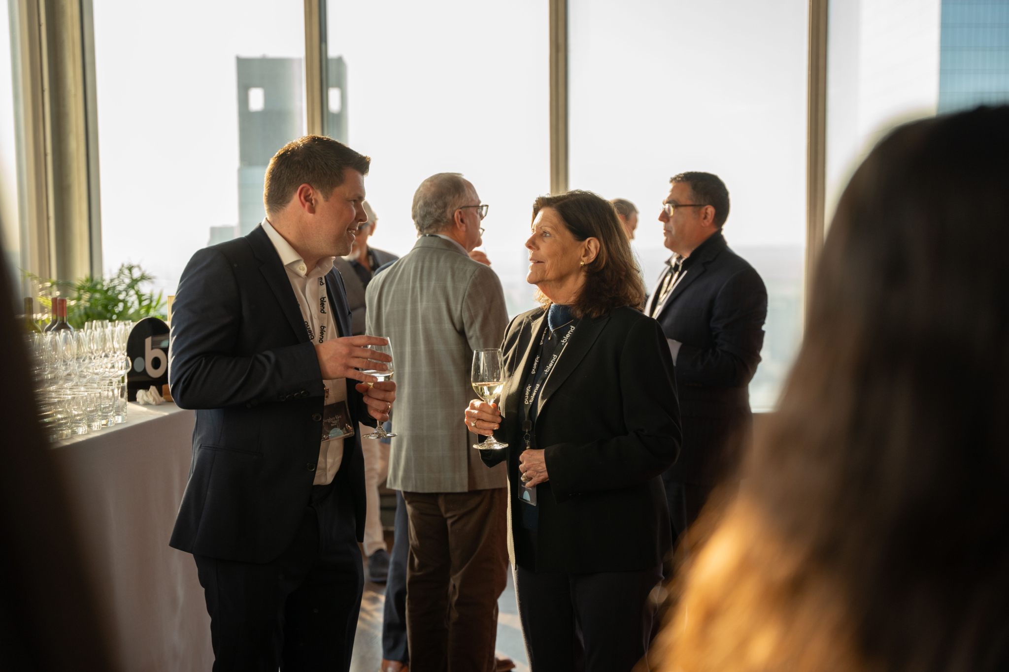 Two C-suite professionals engage in conversation at a Blend community networking event. They wear Blend lanyards and hold glasses of wine.