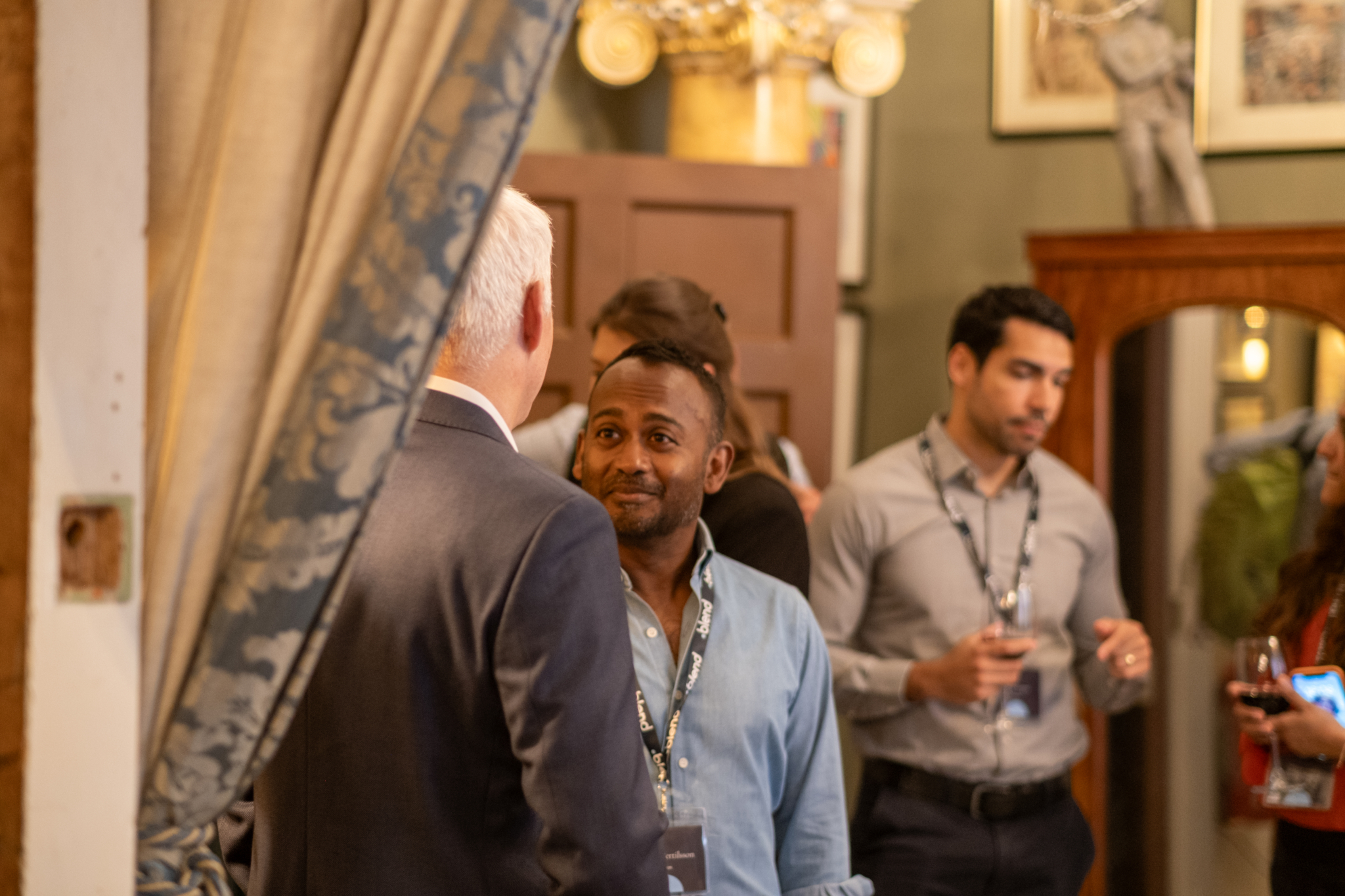 A group of business-wear dressed C-suite professionals talk at a Blend community networking event, wearing Blend branded lanyards.