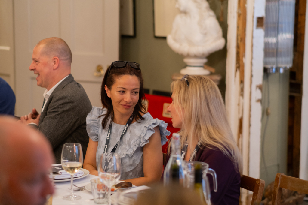 Two C-suite professionals talk at a Blend community networking event, wearing Blend branded lanyards. They are seated at a long dining table.